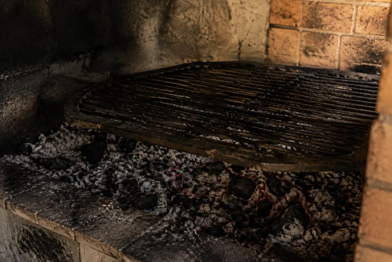 Braseiro pronto dentro da churrasqueira de alvenaria, com o carvão coberto de cinza cinzenta e brasa alaranjada por baixo, sem chama, e a grelha vazia bem acima dele.