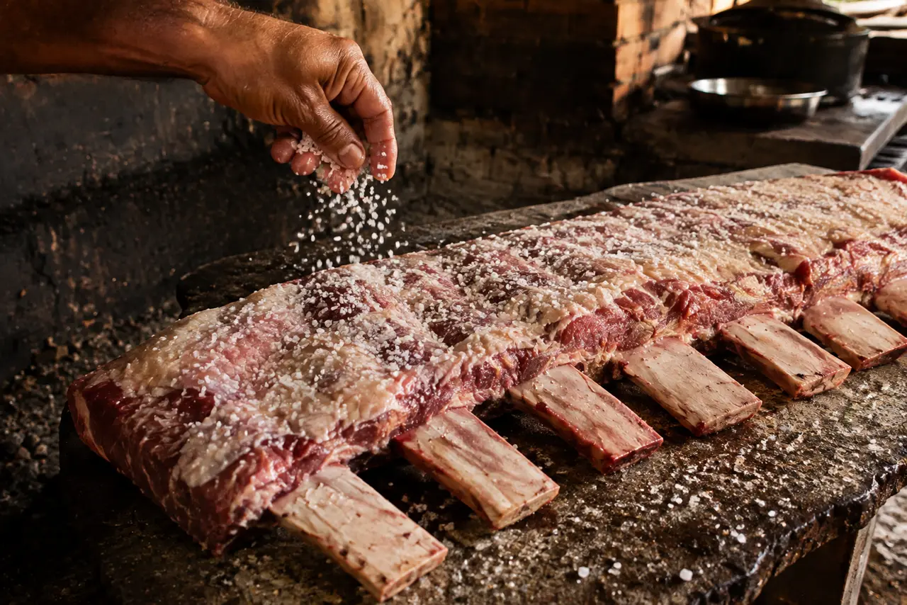 Mão espalhando sal grosso sobre a carne crua da manta de costela, com os grãos brancos visíveis no ar e sobre a superfície.