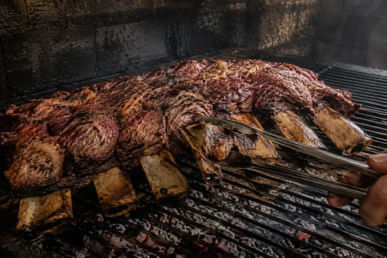 Pinça de churrasco girando um osso central da costela já assada, com o osso quase solto e a carne escura cedendo em volta dele.